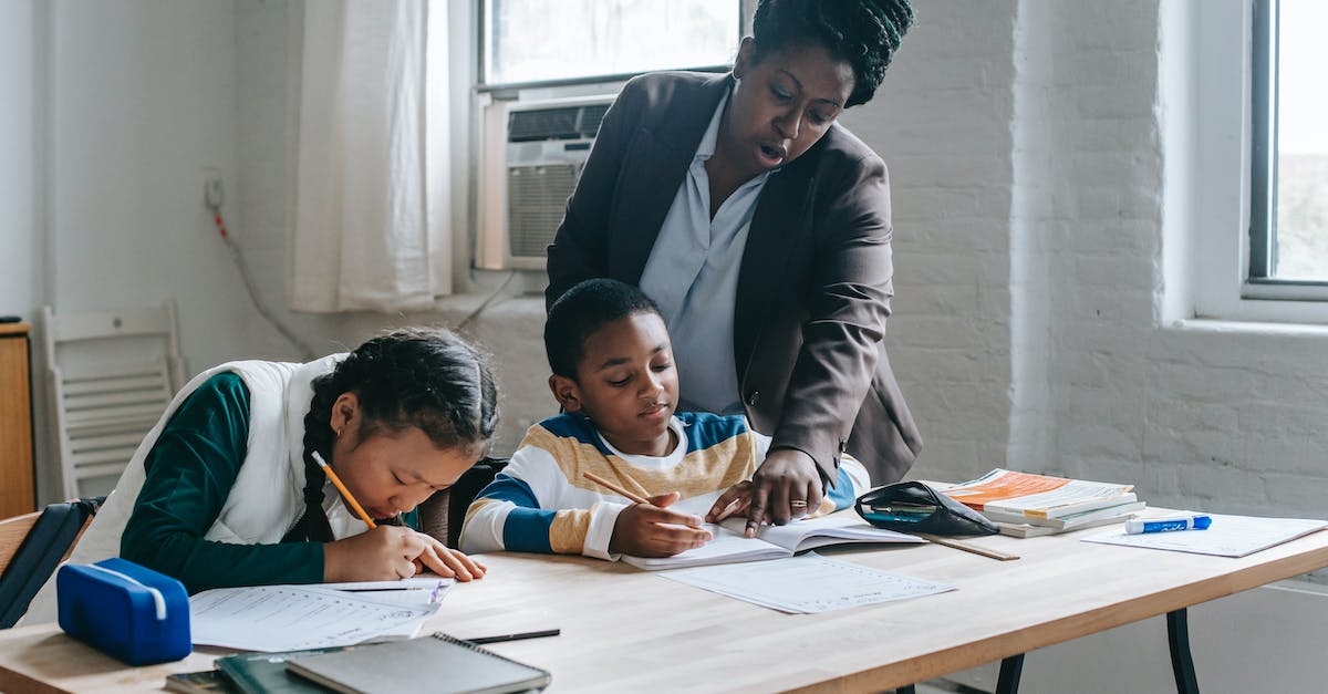 What's the point of Assignments? - Focused African American female teacher in formal clothes standing near school desk with diverse pupils and pointing at schoolboys copybook