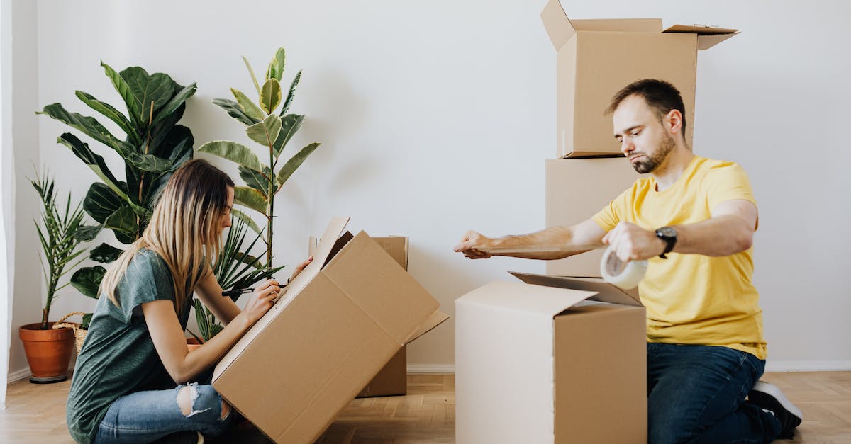 What happens when my box is full? - Side view full body unemotional couple wearing casual clothes and sitting on parquet while sealing carton boxes with tape and writing notes during moving in together
