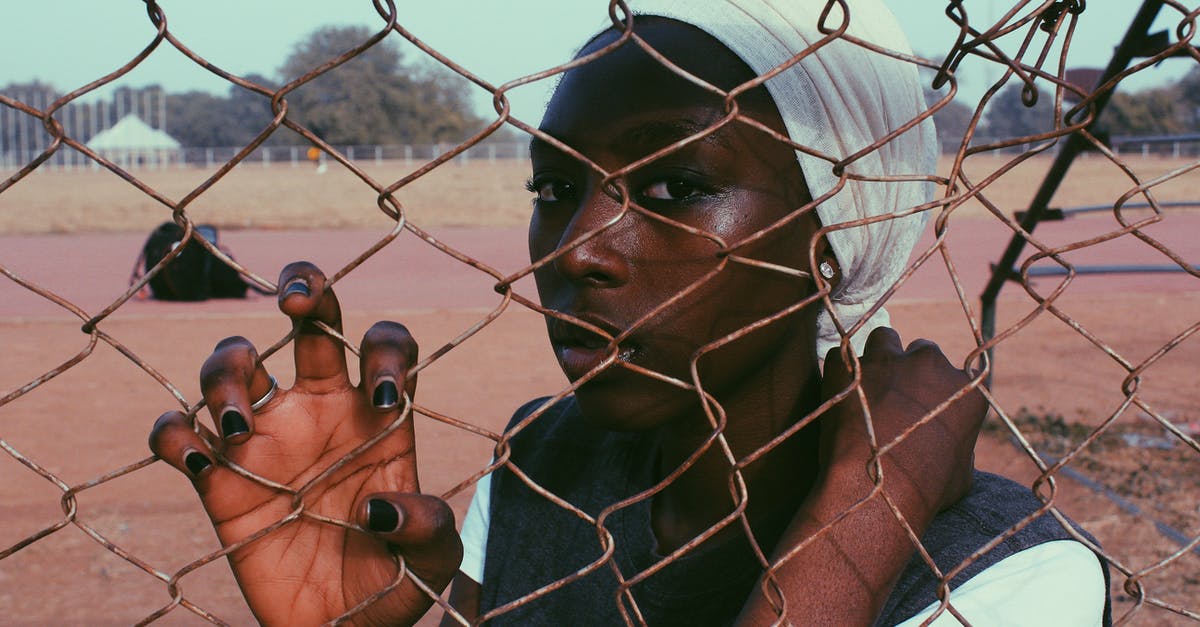 What is behind the mysterious door? - Black thoughtful woman wearing white turban standing behind wire fence on sports ground looking at camera