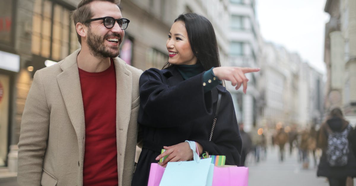 What is the point of investing in shops? - From below happy stylish Asian woman in warm clothes smiling at cheerful unshaven man in glasses and pointing finger away while walking along street together in city after shopping