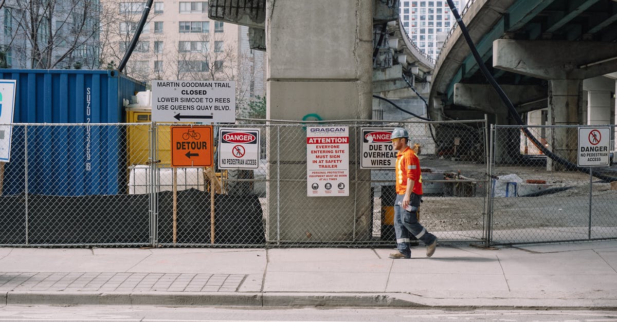 What is this downtown side job during the Corpo prologue? - Full body side view of adult male worker in orange uniform and grey helmet walking under big bridge along chain link fence on street in daylight