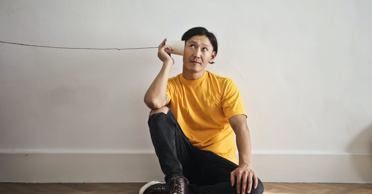 What is this game called? - Asian guy in casual clothes using paper cup with thread as telephone while sitting on floor against white wall