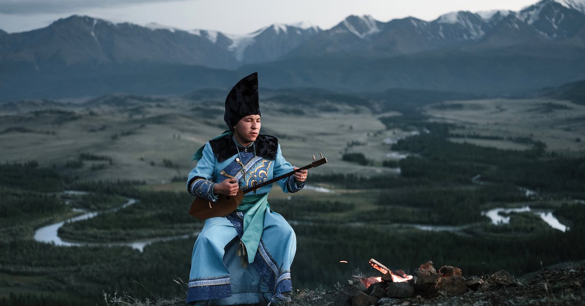 What Melodies can i play to my Mountain? - Full body of concentrated Mongolian male musician in national costume sitting near bonfire and playing traditional musical instrument against picturesque mountains