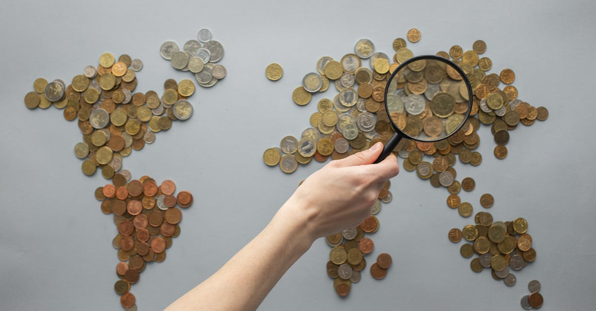 What to do when travelling between countries? - Top view of crop unrecognizable traveler with magnifying glass standing over world map made of various coins on gray background