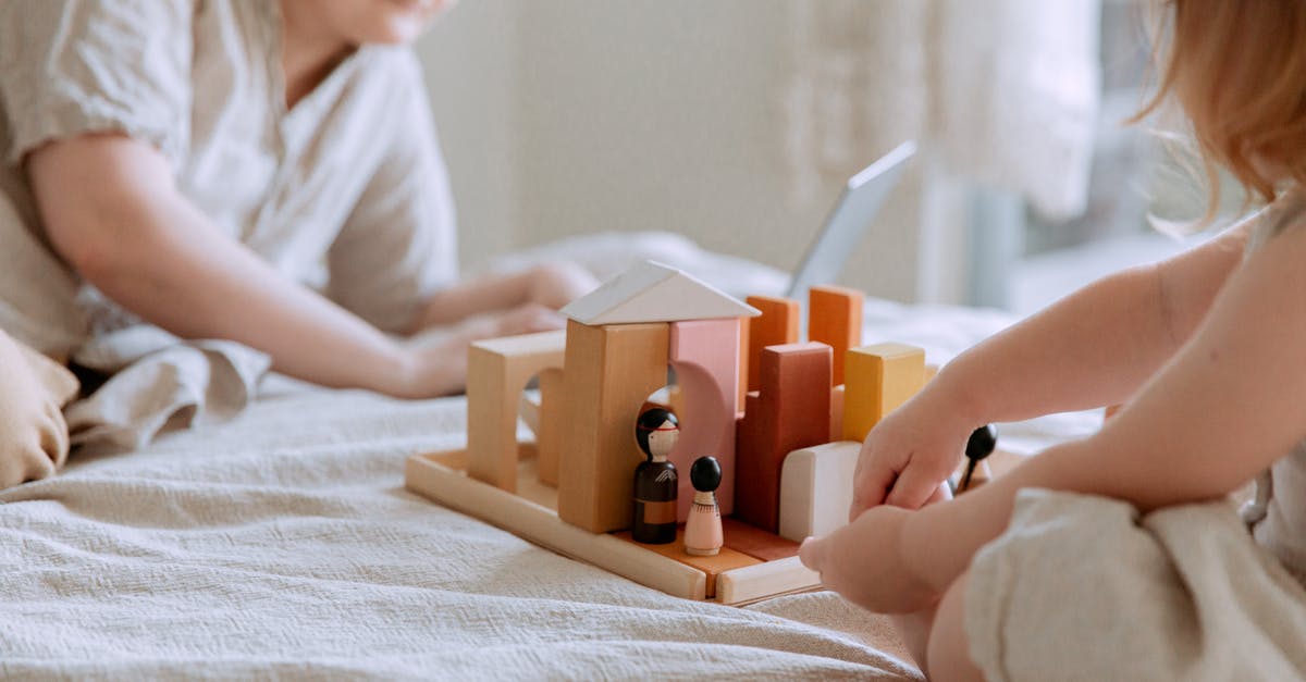 When I break blocks they just reappear? - Faceless toddler girl sitting on bed and playing with wooden blocks and toys while mother using laptop