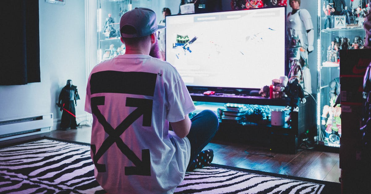 When i play video games on full screen i get a bit of a problem - Back view of anonymous geek male in casual clothes and cap sitting on carpet and playing console video game in living room full of toys and stuff from comic