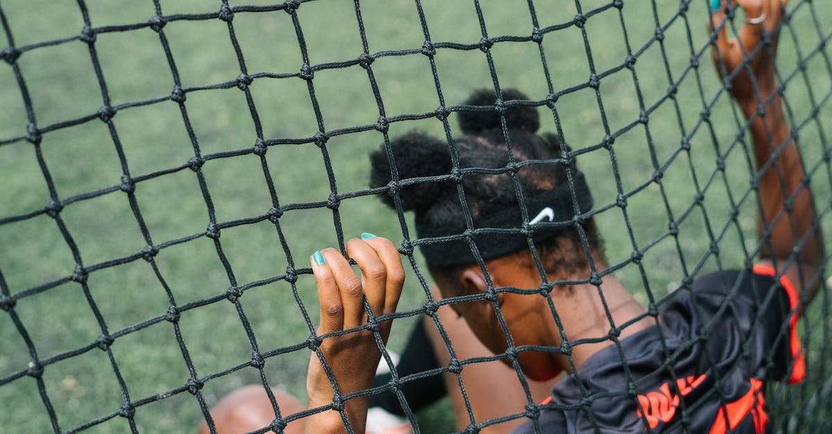 When should I start playing Arena? - Back view of African American teenage football player sitting on field ground and holding grid of gate while preparing to start play football match