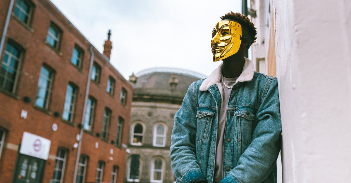 When two dragons fight over a base to the buildings below them get damaged? - From below of confident black activist in stylish outfit and famous Anonymous mask leaning on building placed in city district in daytime