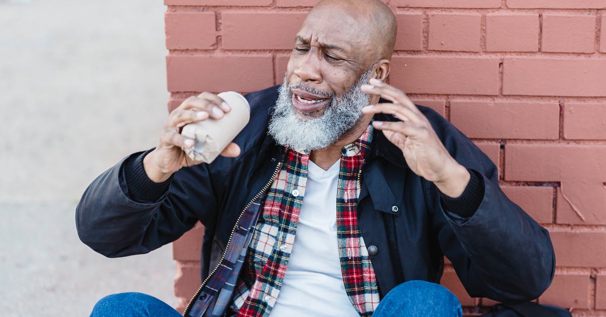 Where can I find a brick? - African American bearded male in casual outfit raising arms with beer can while resting on street near brick wall