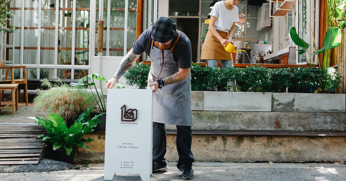 Where do I put the CiaoCiao figurine? - Couple of coworkers in aprons and casual clothes watering plants and putting signboard near cafeteria in daytime