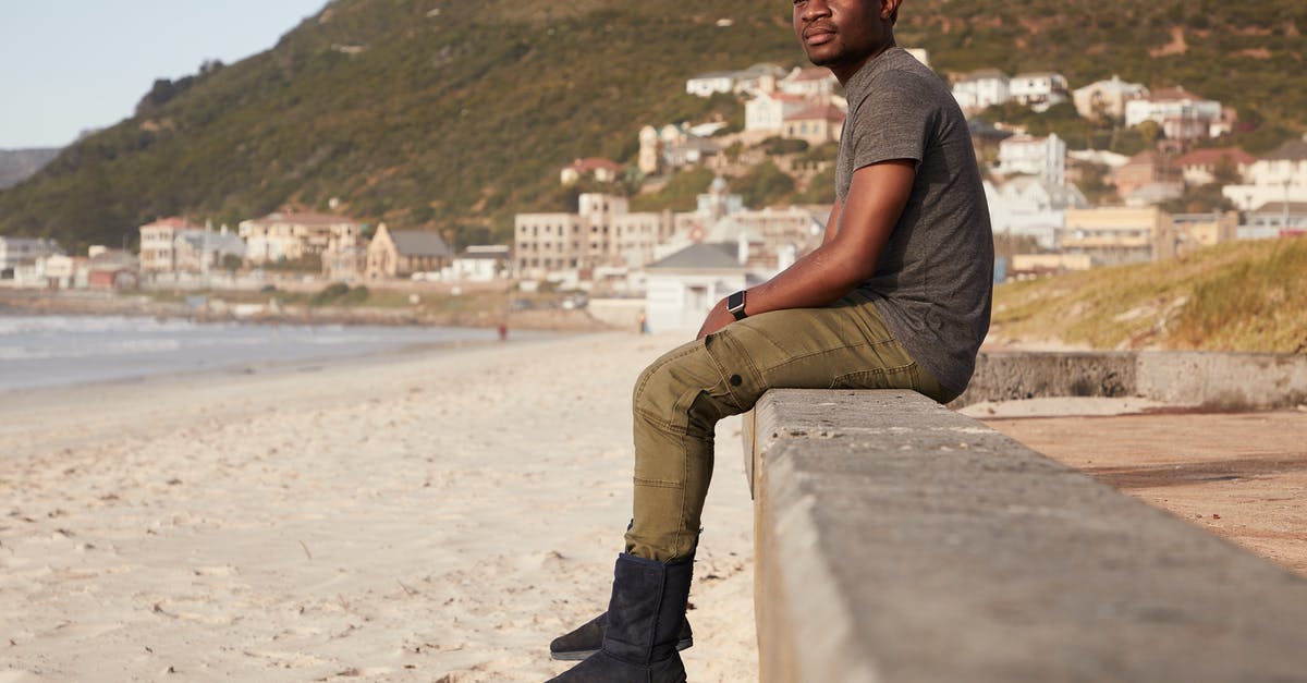 Who is this NPC swimming outside of town borders? - Full body of thoughtful African American male in hat sitting on stone border on sandy coast near sea with coastal town in distance