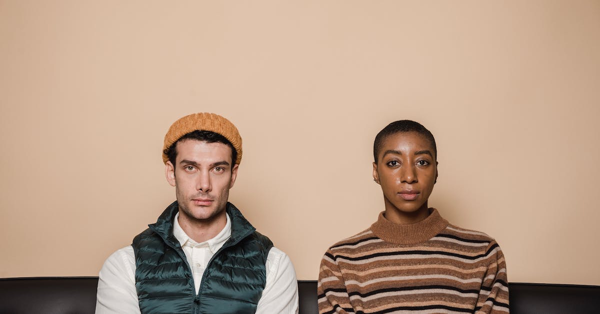 Whoa, A Bad Relationship - Emotionless multiethnic couple sitting on couch against beige background and looking at camera Whoa, A Bad Relationship - Emotionless multiethnic couple sitting on couch against beige background and looking at camera