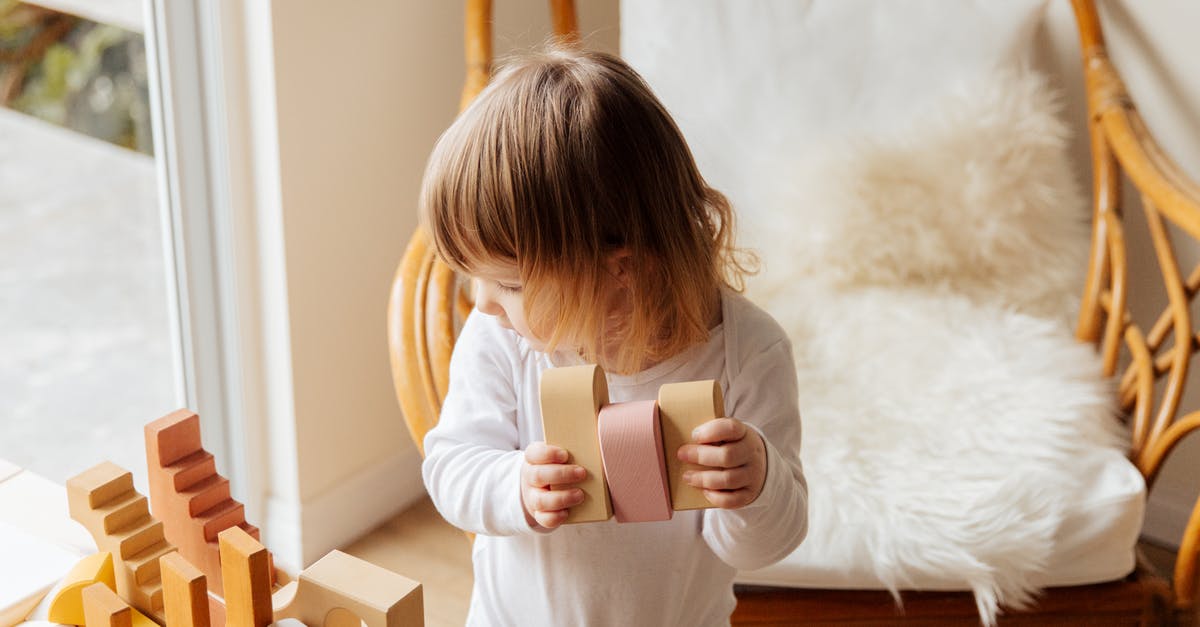 Why build a large spaceship when a small one will do? - From above of cute little child in white wear holding wood blocks for building while standing near wicker armchair