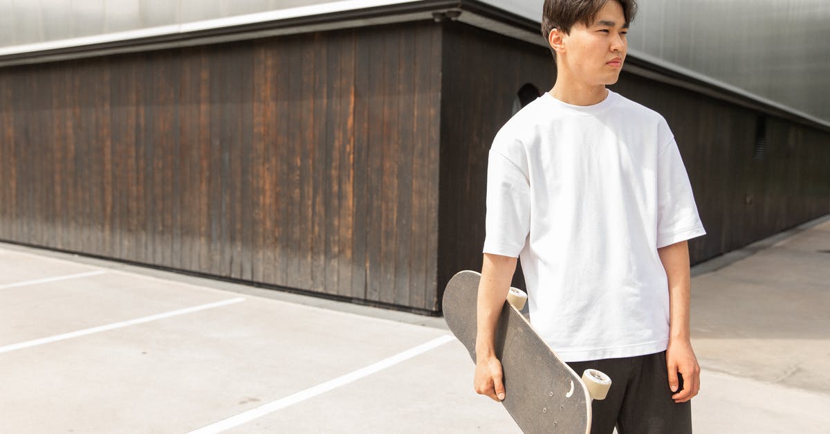 Why can't I build a city here? - Handsome young ethnic guy in white t shirt holding skateboard in hand and looking away thoughtfully while resting near modern building after ride