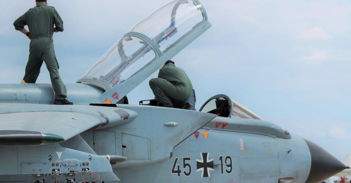 Why do Captain Aldis' men attack me on sight? - Low angle of unrecognizable male pilots in uniforms standing on aged gray multirole combat aircraft before flight against cloudy sunset sky