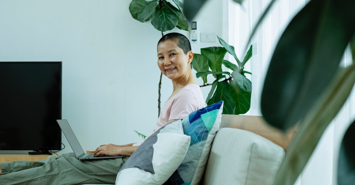 Why does connecting a laptop to a TV via HDMI cause a framerate drop? - Side view of positive middle age ethnic female in casual outfit using laptop on sofa next to plants and window with curtains near TV in house and looking at camera