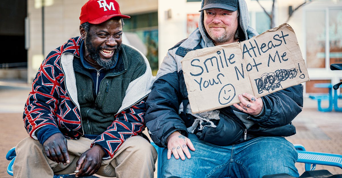 Why does my friend have fewer Pokémon than me? - Positive middle aged man showing carton with smile at least your not me inscription while sitting on bench with laughing African American male friend
