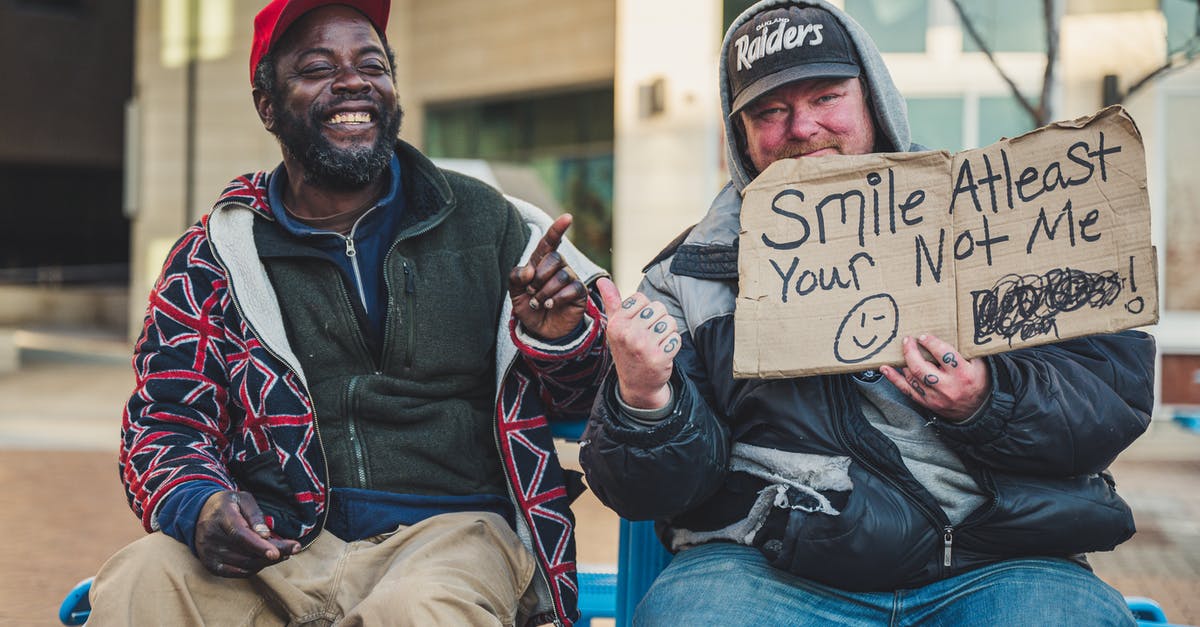 Why does Voorhees call me "china"? - Cheerful black man in cap sitting near plump tattooed male with SMILE AT LEAST YOUR NOT ME inscription on cardboard while showing with fingers at each other on street