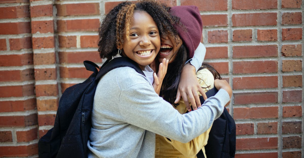 Why don't I have a hat anymore? - Cheerful African American girl in casual clothes hugging friend while having fun near brick wall