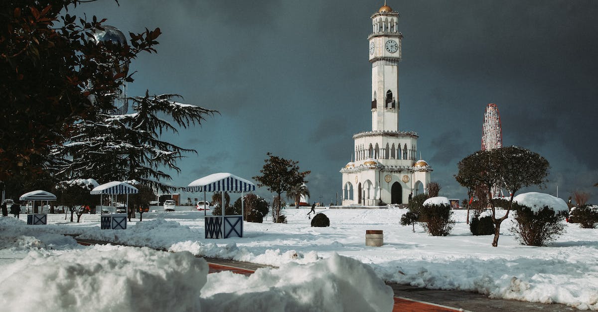 Why is the clock frozen? - Tall white clock tower on snowy terrain near wooden structures and green trees located against overcast sky in Georgia in Batumi city in winter time